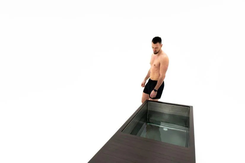 A shirtless man in black shorts stands beside a rectangular glass tank filled with water, reminiscent of a Chill Tubs - Professional affordable Ice Baths ideal for gym fanatics. The background is entirely white, giving the scene a minimalist appearance. The man is looking down at the water inside the tank.