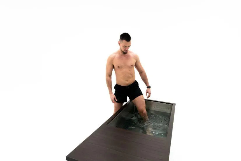 A shirtless man wearing black shorts is standing with one leg submerged in a Chill Tubs - Professional affordable Ice Baths ideal for gym fanatics, a rectangular water tank with a wooden edge designed for cold water therapy. The background is plain white.