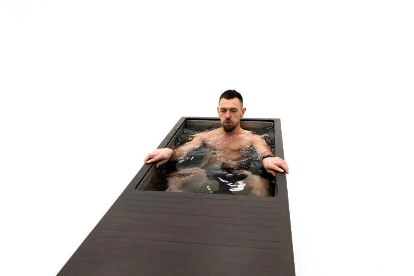 A man is submerged in a sleek, narrow black Chill Tubs - Professional affordable Ice Baths ideal for gym fanatics by Chill Tubs filled with water, possibly indulging in cold water therapy, with his arms resting on the sides. The background is completely white, creating a minimalist and modern contrast.