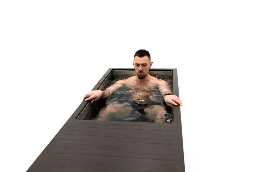 A man with dark hair and a beard is sitting in a modern, rectangular black tub filled with water. He appears relaxed with his eyes slightly closed, holding the edges of the tub. This setting is part of his cold water therapy routine using a Chill Tubs - Professional affordable Ice Baths ideal for gym fanatics by Chill Tubs. The background is plain white.