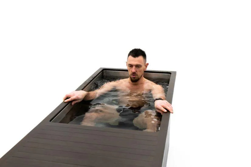 A man with short dark hair is sitting in a modern rectangular Chill Tubs - Professional affordable Ice Baths ideal for gym fanatics filled with water. He is looking down and has his arms resting on the sides of the tub, perhaps engaging in cold water therapy. The background is completely white.