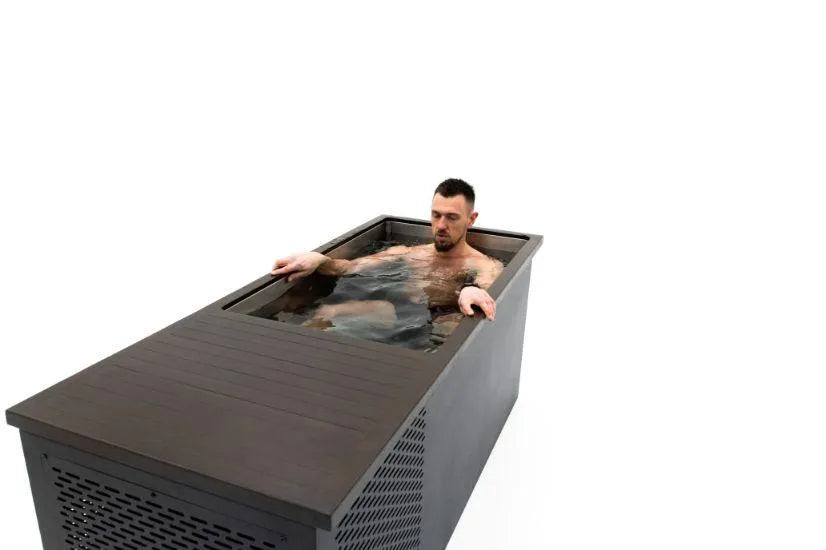 A man with a beard sits in a modern, rectangular Chill Tubs - Professional affordable Ice Baths ideal for gym fanatics by Chill Tubs, filled with cold water, against a plain white background. The front of the tub has a grated panel. Submerged to his chest, arms resting on the sides, he appears calm and focused as he embraces cold water therapy.