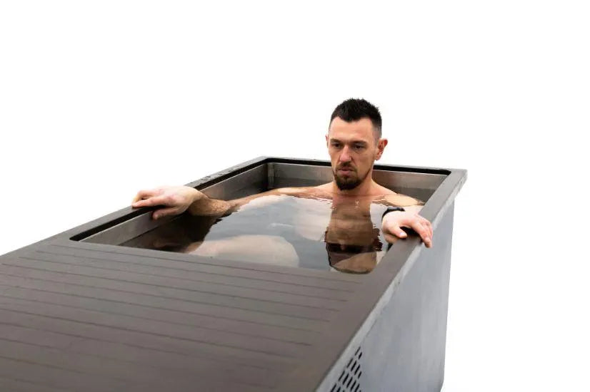 A man with short dark hair and a beard sits in a rectangular Chill Tubs - Professional affordable Ice Baths ideal for gym fanatics filled with water, practicing cold water therapy. He has a neutral expression, one hand resting on the Chill Tubs' edge, and the other arm submerged. The background is plain and white.