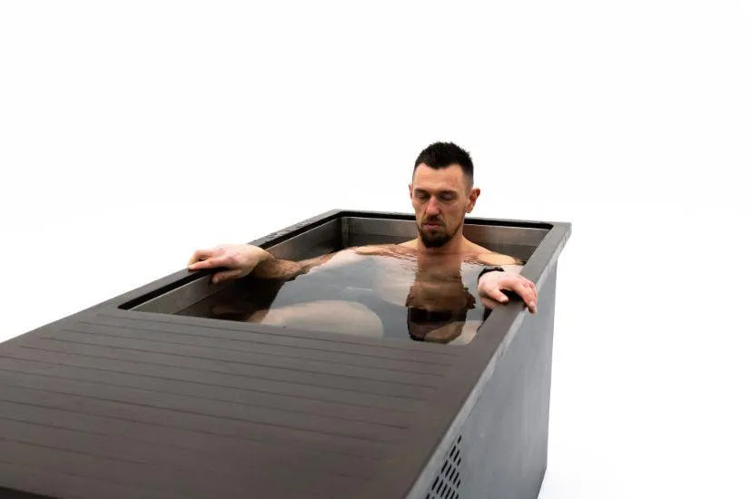 A man with short dark hair and a beard, wearing a wrist monitor on his left wrist, is sitting in a Chill Tubs - Professional affordable Ice Baths ideal for gym fanatics. He looks focused and calm, with his eyes closed and his right hand resting on the edge of the bath. The background is plain white.