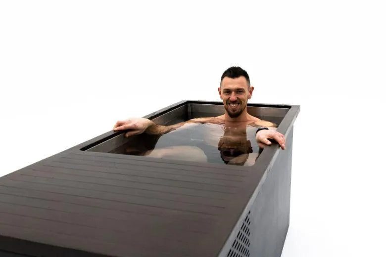 A man with short dark hair and a beard is sitting and smiling in a dark-colored bathtub filled with cold water. The background is completely white, making the tub and the man the focal points of the image, perfect for showcasing Chill Tubs - Professional affordable Ice Baths ideal for gym fanatics by Chill Tubs' cold water therapy benefits.