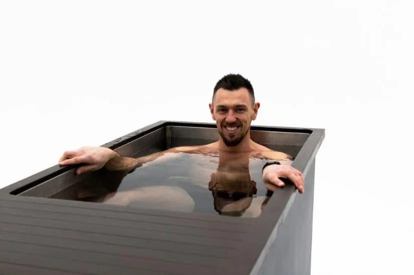 A man with short dark hair and a beard is smiling while sitting in a rectangular metal tub filled with water, practicing cold water therapy using the Chill Tubs - Professional affordable Ice Baths ideal for gym fanatics from Chill Tubs. The background is plain white. He has one arm resting on the side of the tub and wears a black wristband.