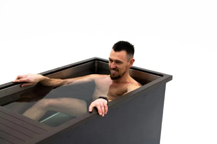 A man with a short beard and short hair, wearing swim trunks and a watch, is sitting in a rectangular black tub filled with water. He is smiling and resting one arm on the side of the tub against a plain white background, enjoying some cold water therapy in his Chill Tubs - Professional affordable Ice Baths ideal for gym fanatics by Chill Tubs.