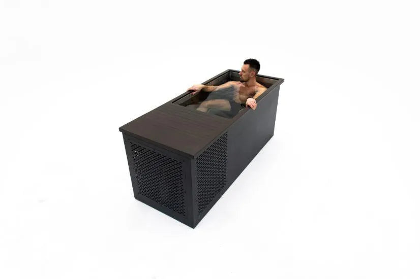 A man is relaxing in a modern, rectangular wooden bathtub filled with water, indulging in cold water therapy. The minimalist design of the Chill Tubs - Professional affordable Ice Baths ideal for gym fanatics features clean lines and a dark finish, set against a plain, white background.