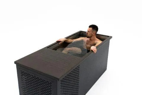 A man with short dark hair and a beard is sitting in a rectangular black tub filled with water, looking to his left. The tub, a Chill Tubs - Professional affordable Ice Baths ideal for gym fanatics from Chill Tubs, is placed in a white space, and the man's upper body is visible above the water. He's engaged in cold water therapy as part of his routine.
