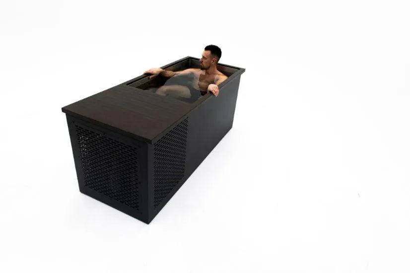 A person is sitting in a modern, rectangular wooden tub filled with water. The Chill Tubs - Professional affordable Ice Baths ideal for gym fanatics, designed by Chill Tubs for cold water therapy, has a dark finish and features a perforated design on one side. The background is plain white, giving the scene a minimalist look. The person appears relaxed, with arms resting on the tub's edges.