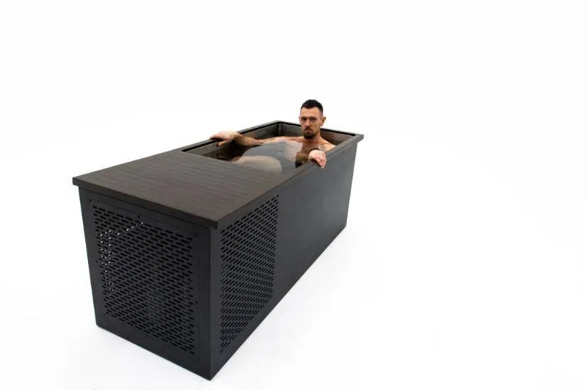 A man sits partially submerged in a modern, rectangular Chill Tubs - Professional affordable Ice Baths ideal for gym fanatics by Chill Tubs with a dark finish. The tub, designed for cold water therapy, is placed against a plain white background, highlighting its sleek design and lattice-style ventilation panel on one side.