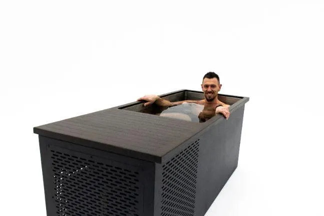 A man is relaxing in a modern, dark-colored wooden bathtub filled with water. The bathtub, known as Chill Tubs - Professional affordable Ice Baths ideal for gym fanatics, has a perforated side panel and a rectangular shape. He appears content and is smiling while enjoying the benefits of cold water therapy. The background is plain white.