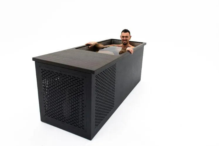 A man is sitting in a modern, rectangular wooden bathtub filled with cold water. The sleek, dark-finished tub stands alone on a white background, resembling the Chill Tubs - Professional affordable Ice Baths ideal for gym fanatics used for cold water therapy. The man is smiling, with his shoulders and head visible above the water.