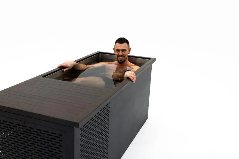 A person with short dark hair and a beard is relaxing in a rectangular dark-colored Chill Tubs - Professional affordable Ice Baths ideal for gym fanatics filled with water. The tub has a modern design with slatted and solid panels. The background is completely white. The person looks cheerful and content.