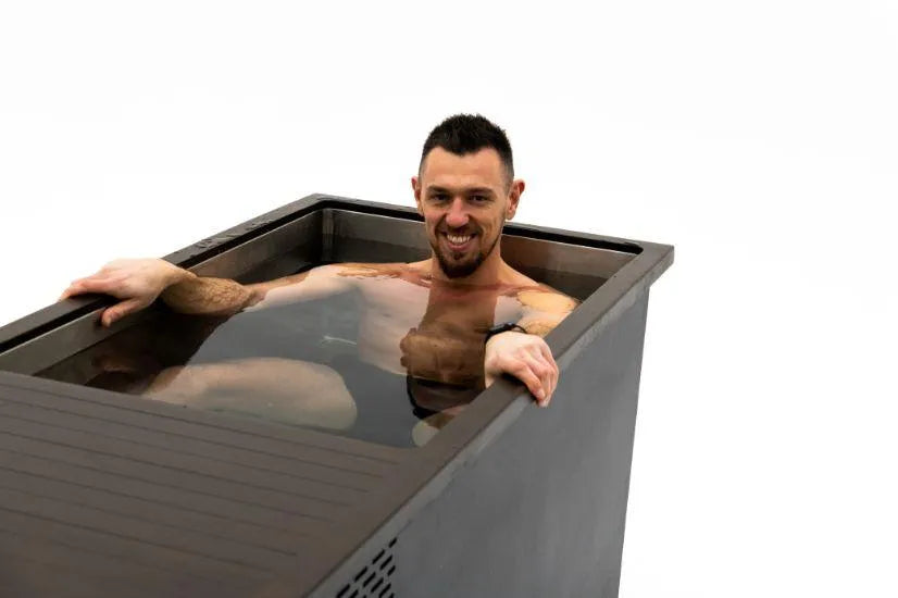 A man with short dark hair and a beard is smiling while sitting in a large black rectangular Chill Tubs - Professional affordable Ice Baths ideal for gym fanatics, partially submerged in water. He is shirtless, showcasing tattoos on his arm, enjoying his cold water therapy session. The background is plain and white.