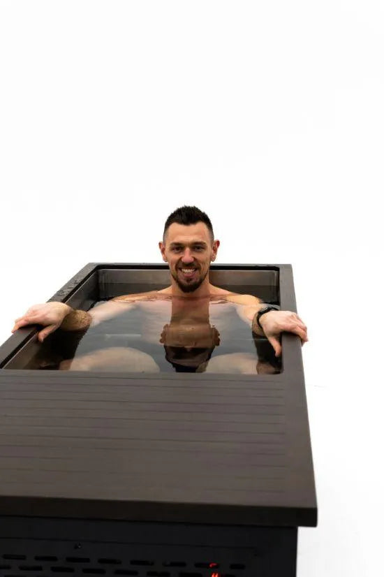 A man with short dark hair and a beard is smiling as he relaxes in a modern, rectangular bathtub filled with cold water. Both his arms rest on the edges of the tub, enjoying some Chill Tubs - Professional affordable Ice Baths ideal for gym fanatics therapy against a plain white background.