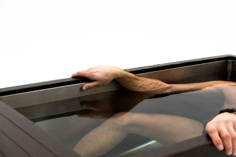A person partially submerged in a Chill Tubs - Professional affordable Ice Baths ideal for gym fanatics filled with water up to the chest. One arm is resting on the edge of the tank. The person appears to have short hair and is possibly undergoing cold water therapy or relaxation treatment. The background is plain white.