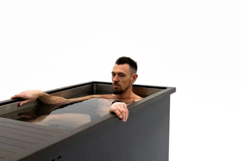 A man with facial hair sits immersed up to his chest in a sleek, rectangular black metal Chill Tubs - Professional affordable Ice Baths ideal for gym fanatics filled with water against a plain white background. He appears calm and relaxed, resting one arm on the edge of the tub, enjoying the benefits of cold water therapy from Chill Tubs.