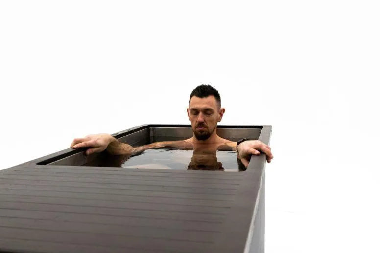 A man with short dark hair and a beard is sitting and soaking in a rectangular, modern dark wood Chill Tubs - Professional affordable Ice Baths ideal for gym fanatics filled with water. The background is stark white, making the man and the Chill Tubs the focal points of the image.