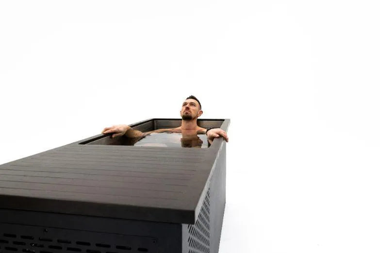 A man with dark hair and a beard is submerged up to his chest in a rectangular black sensory deprivation tank, also known as an isolation tank, filled with water. The background is completely white, highlighting the individual and the tank, which could be mistaken for one of those Chill Tubs - Professional affordable Ice Baths ideal for gym fanatics from Chill Tubs.