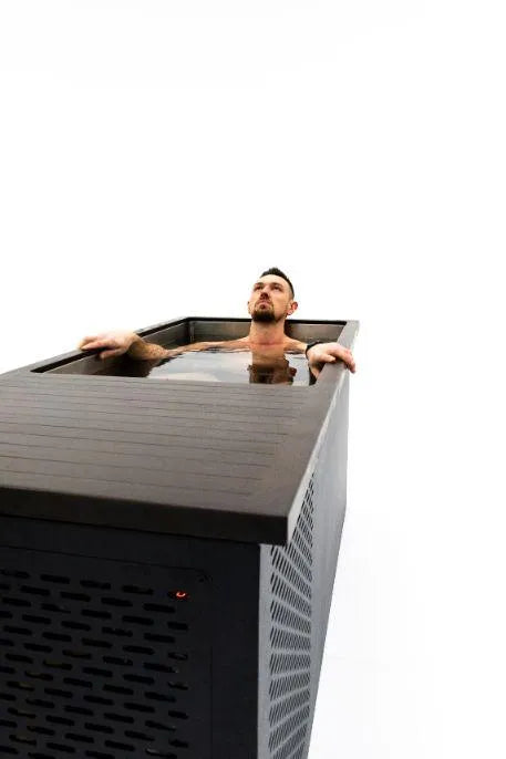 A person with a trimmed beard and short hair is relaxing in a black rectangular Chill Tubs - Professional affordable Ice Baths ideal for gym fanatics filled with water. Their arms rest on the sides of the tub, and they appear to gaze thoughtfully ahead, perhaps savoring the benefits of cold water therapy. The background is white and minimalistic.