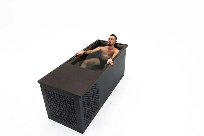 A shirtless man with short hair is lying in a modern dark bathtub filled with water, designed to resemble a Chill Tubs - Professional affordable Ice Baths ideal for gym fanatics. The tub has a rectangular shape with an external wooden texture and slatted design on one side. The background is completely white.