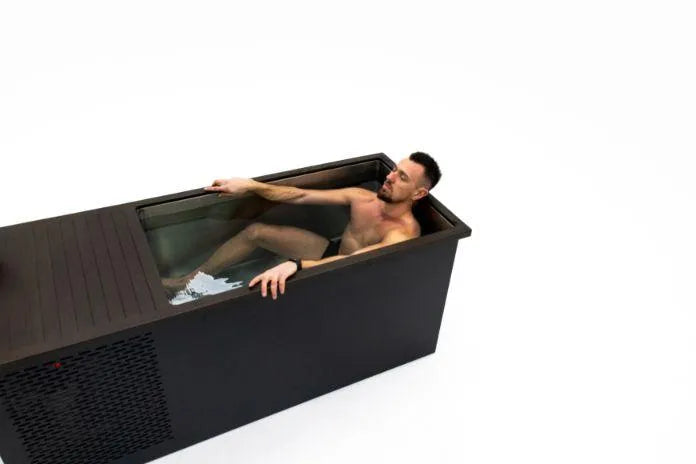 A man relaxes in a sleek, rectangular Chill Tubs - Professional affordable Ice Baths ideal for gym fanatics, partially covered with a wooden lid. He is sitting back, immersed in the frigid water for cold water therapy, with one arm resting on the edge. The background is plain white, highlighting the bath setup.