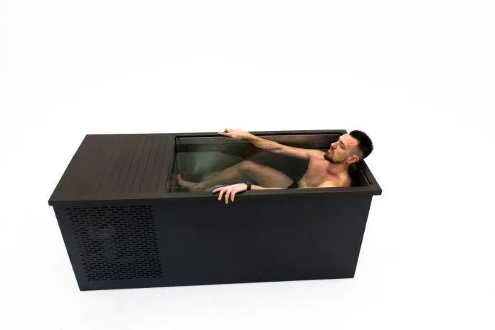 A man is taking an ice bath in a rectangular black Chill Tubs - Professional affordable Ice Baths ideal for gym fanatics from Chill Tubs. Wearing black shorts, he is partially submerged in water and appears to be adjusting something on the far side of the tub. The background is plain white, emphasizing his focus on cold water therapy.