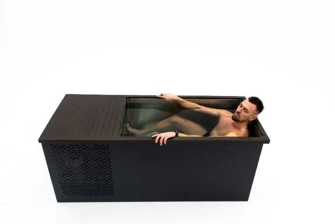A person is submerged in a compact black Chill Tubs - Professional affordable Ice Baths ideal for gym fanatics, with a lid partially slid open. The individual appears to be relaxing, enjoying some cold water therapy, with one arm resting on the side and the other adjusting the lid. The background is plain white.