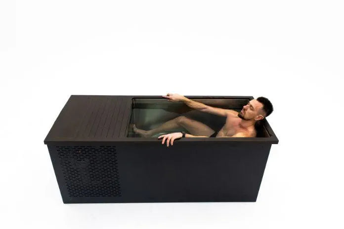 A man is in a minimalist, rectangular Chill Tubs - Professional affordable Ice Baths ideal for gym fanatics with a black exterior. The tub has a cover partially over it, and the man is leaning back, his right arm resting on the edge and left arm submerged in the water. The background is plain white, emphasizing his serene cold water therapy session.