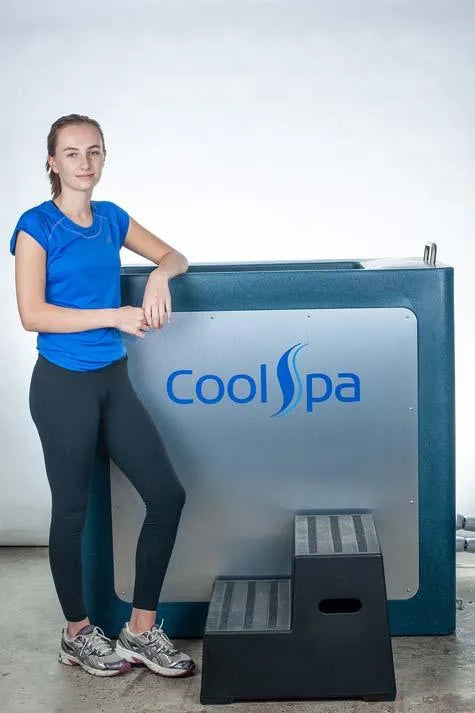 A woman in athletic clothing stands next to a Hot Tub Liverpool CryoHome Ice Bath Spa 1 featuring digital temperature control. She has her arm resting on the chamber and is smiling. The metallic chamber displays a blue "Hot Tub Liverpool" logo, and gray steps are positioned in front of it.
