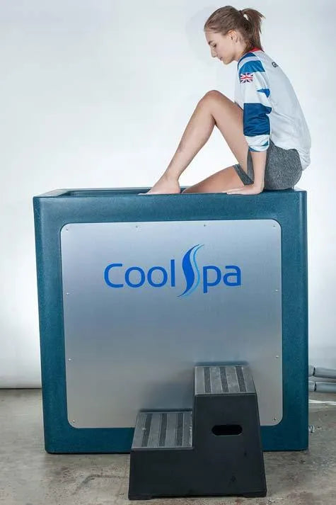 A person in athletic wear is sitting on the edge of a large, square, blue container labeled "Hot Tub Liverpool CryoHome Ice Bath Spa 1." They are dipping their legs into the container, which features digital temperature control for precise cooling. A small set of gray and black steps leads up to the Hot Tub Liverpool chiller kit.