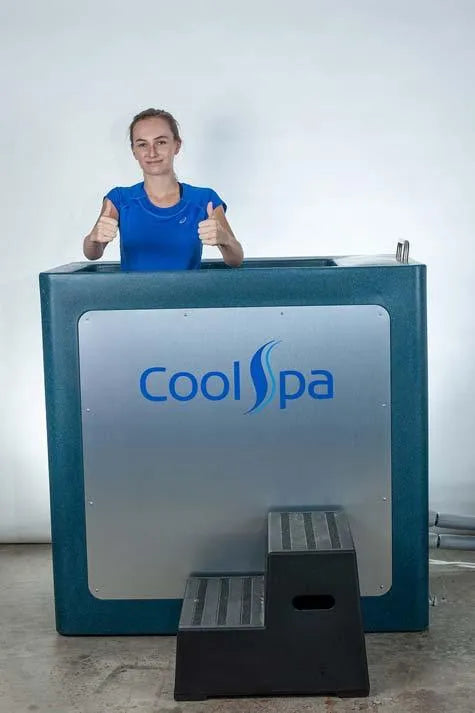 A person standing inside a CryoHome Ice Bath Spa 1, giving two thumbs up. The machine, equipped with digital temperature control and the Hot Tub Liverpool logo on its gray front panel, features a small black step ladder in front. The individual is wearing a blue shirt and appears enthusiastic.