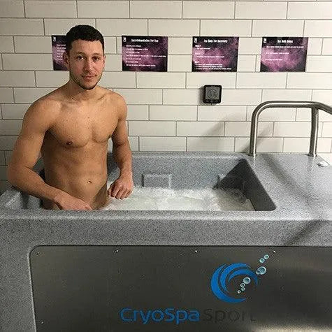 A shirtless man stands in a gray CryoSpa Sport - Cold Water Ice Bath filled with cold water, ideal for athlete recovery. He is positioned against a tiled wall, looking directly at the camera. The CET CryoSpas logo is visible on the front of the bath.