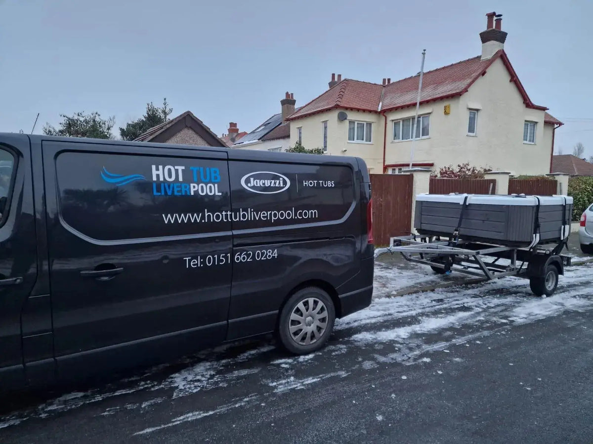 A black van with business advertising for "Hot Tub Liverpool" and a contact number is parked on a snowy residential street in North West England. The van, involved in Hot Tub Relocation Services - Hot Tub Liverpool, is towing a small trailer carrying a large boxed item, likely a hot tub. Snow partially covers the ground and rooftops of nearby houses.