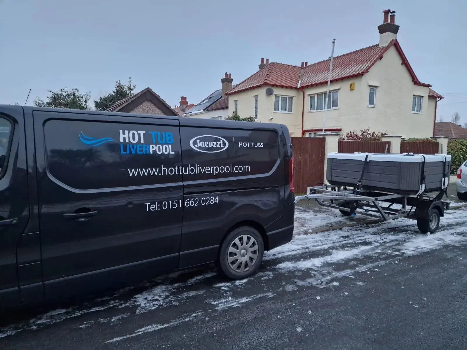 A black van with business advertising for "Hot Tub Liverpool" and a contact number is parked on a snowy residential street in North West England. The van, involved in Hot Tub Relocation Services - Hot Tub Liverpool, is towing a small trailer carrying a large boxed item, likely a hot tub. Snow partially covers the ground and rooftops of nearby houses.