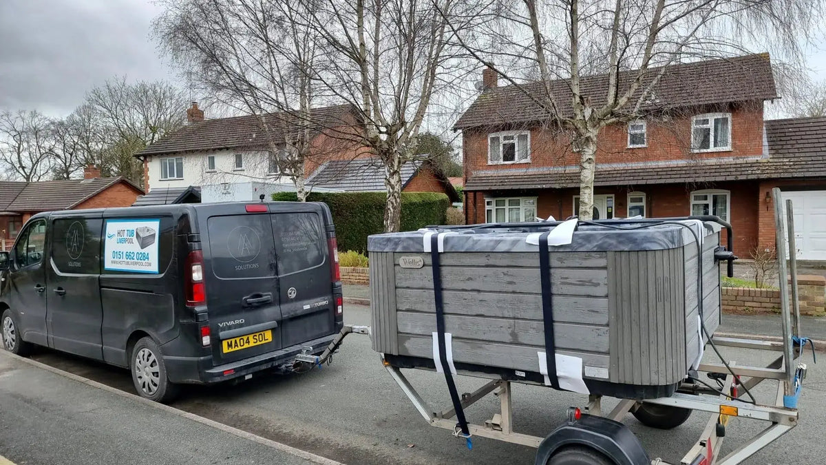 A black van is parked on a residential street in Liverpool, towing a trailer carrying a large hot tub secured with straps. Brick houses and bare trees are in the background. The van, marked with "Hot Tub Liverpool" and a phone number, boasts the license plate "MA04 SJL," indicating its North West England origin.