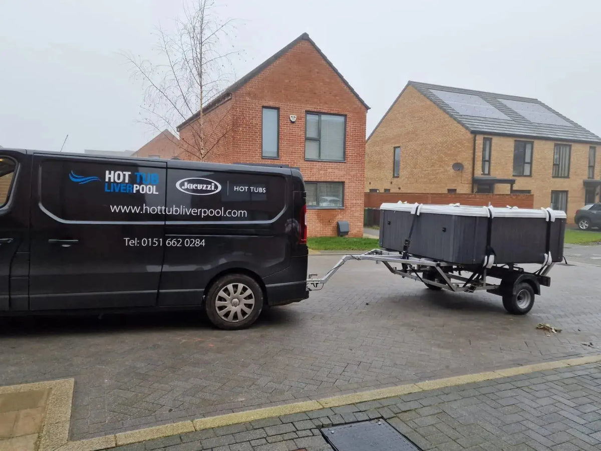 A black van with the text "HOT TUB LIVERPOOL" and "Hot Tub Relocation Services - Hot Tub Liverpool" on its side is parked on a residential street in North West England. It is towing a trailer carrying a hot tub. The background reveals two brick houses with modern architecture, exemplifying a typical hot tub relocation scene.