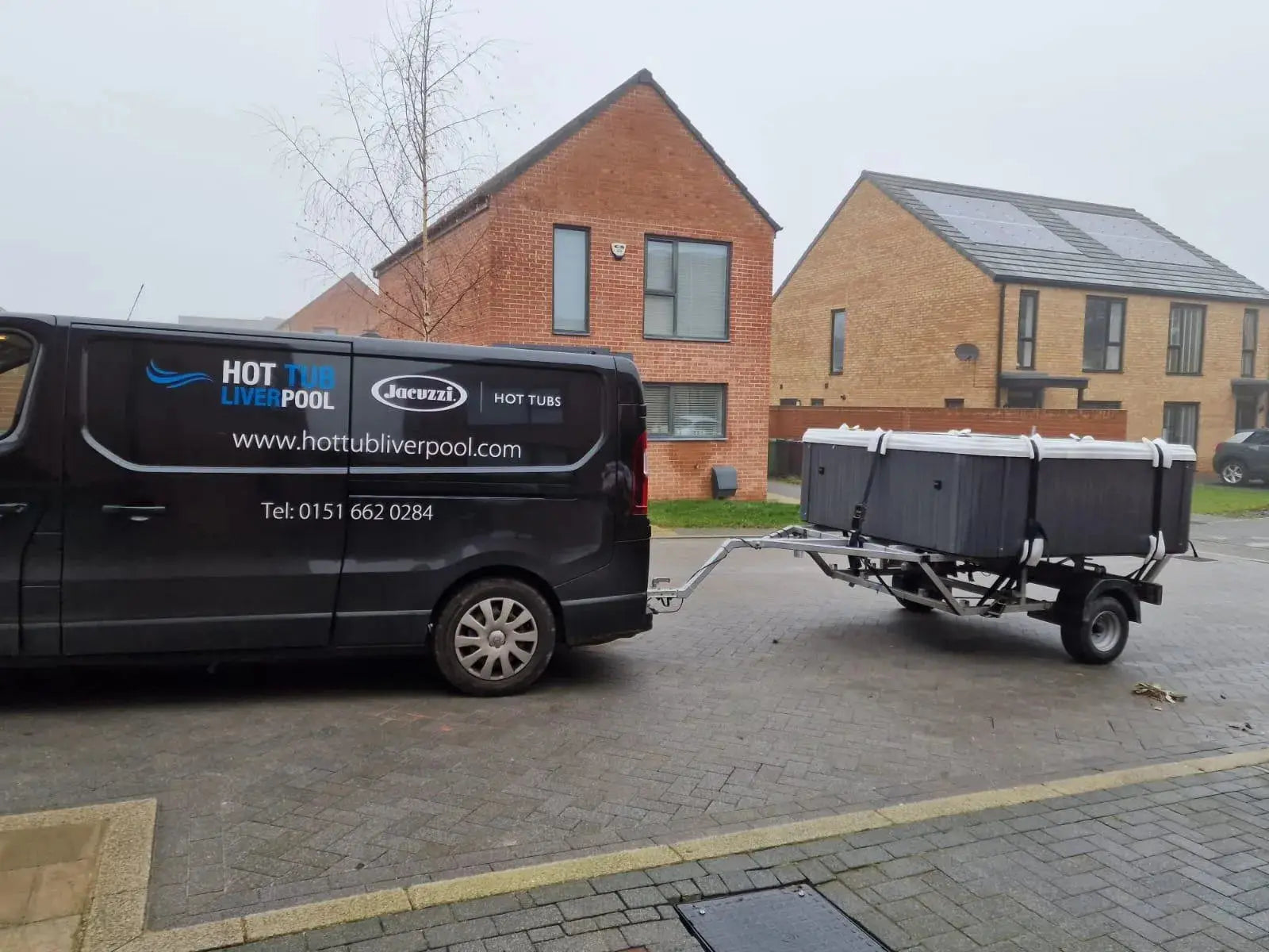 A black van with the text "HOT TUB LIVERPOOL" and "Hot Tub Relocation Services - Hot Tub Liverpool" on its side is parked on a residential street in North West England. It is towing a trailer carrying a hot tub. The background reveals two brick houses with modern architecture, exemplifying a typical hot tub relocation scene.
