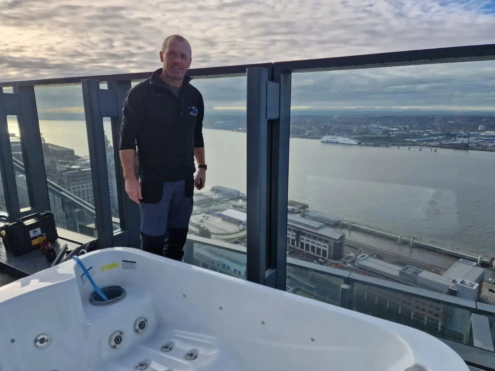A person wearing a dark jacket and shorts is standing on a rooftop terrace in Liverpool beside an empty hot tub. The terrace boasts a glass railing with views of the city's waterline, buildings, and ships in the distance under a cloudy sky, perhaps suggesting an upcoming **Hot Tub Relocation Services - Hot Tub Liverpool** by **Hot Tub Liverpool** in North West England.