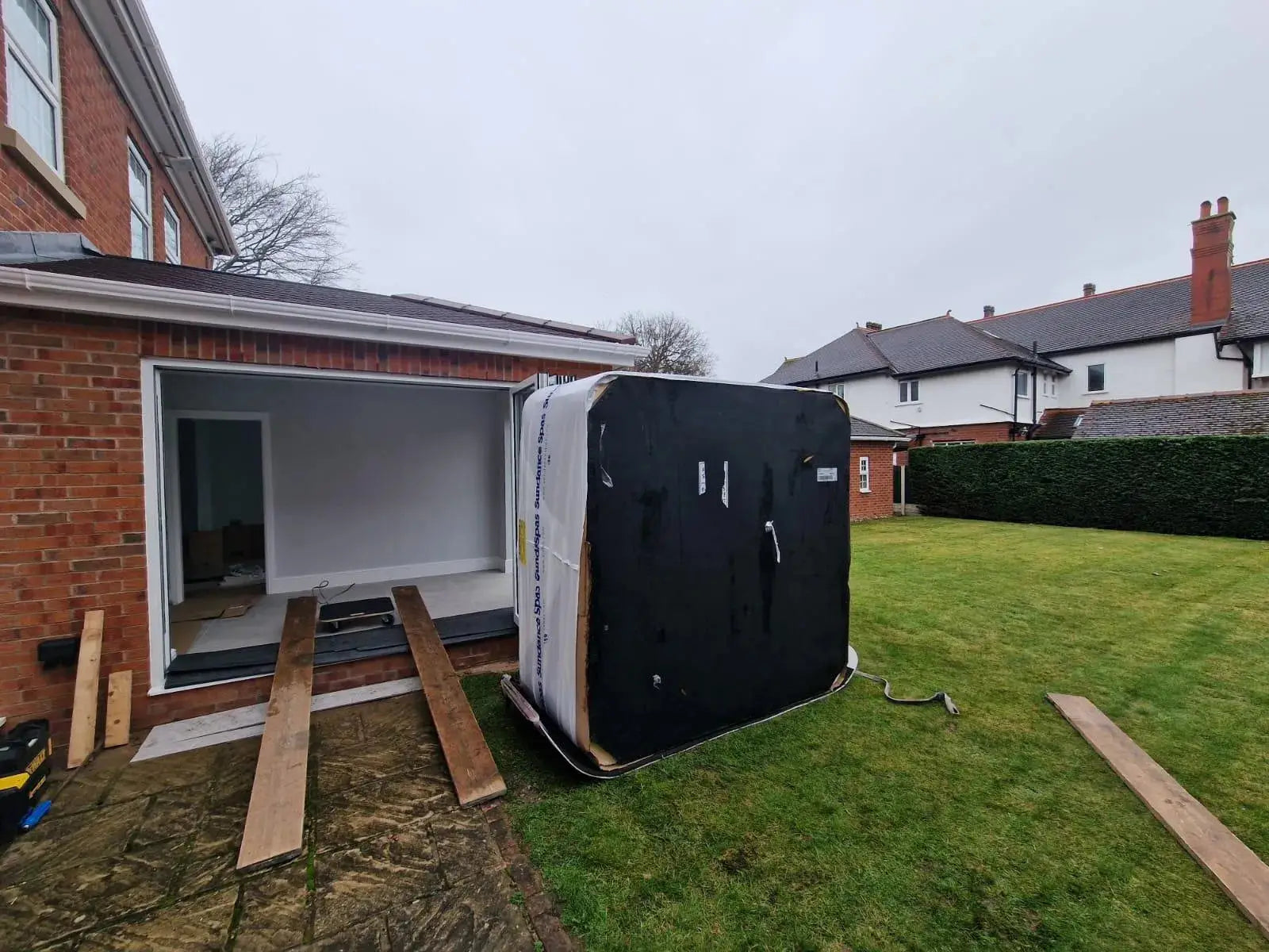A large black and white rectangular object, possibly a hot tub, is being transported through an opening in a brick house. Wooden planks are laid as a ramp leading into the house. The scene includes a grassy yard and neighboring brick houses, reminiscent of suburban Liverpool in North West England. Hot Tub Relocation Services - Hot Tub Liverpool is providing the service for this move.