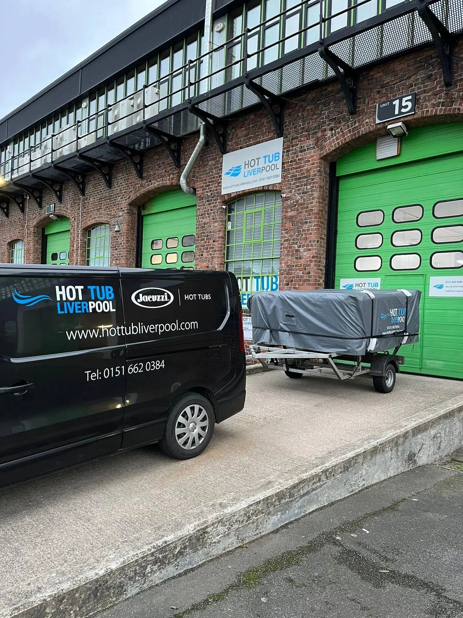 A black van labeled "Hot Tub Relocation Services - Hot Tub Liverpool" is parked outside a brick building with a green garage door, also labeled "Hot Tub Relocation Services - Hot Tub Liverpool." A trailer next to the van carries a covered item, likely for hot tub relocation. The building, located in North West England, has a sign with "15" above the door.