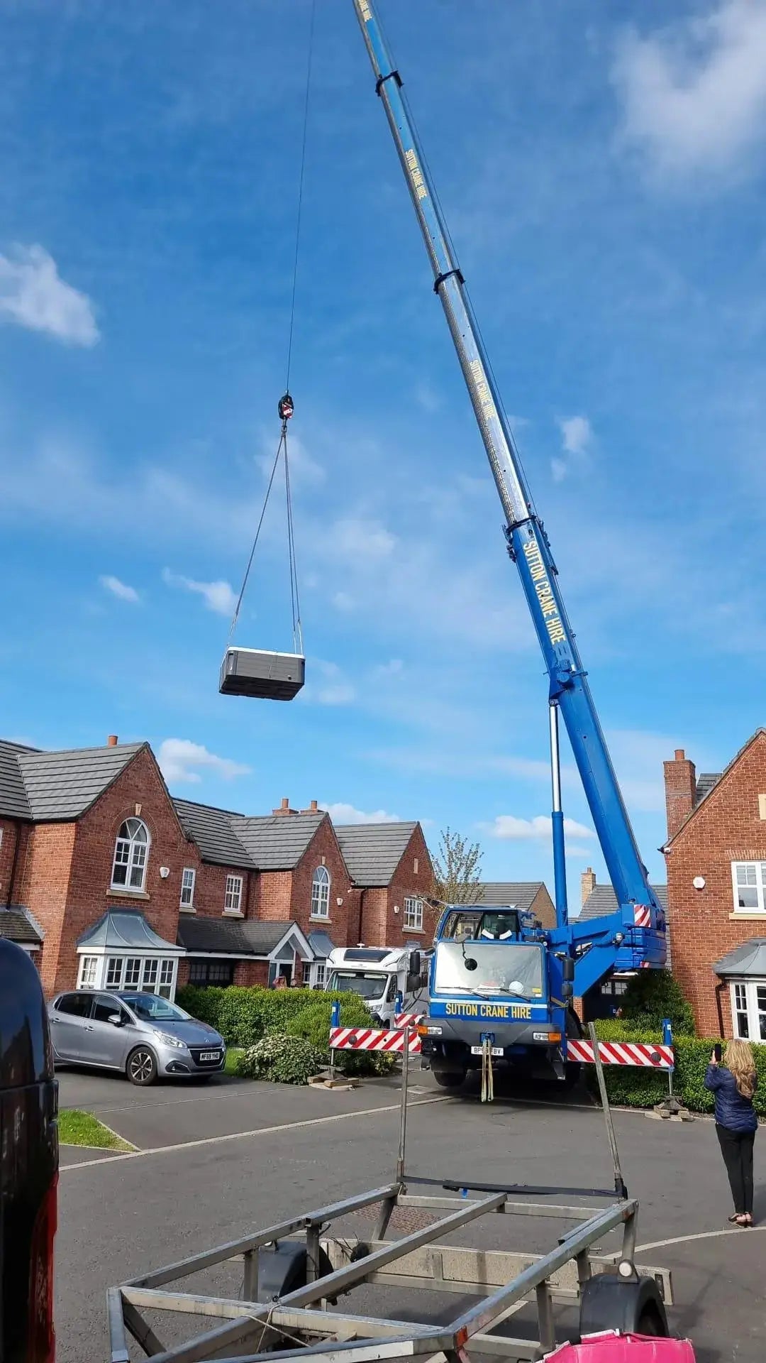 A large blue crane is lifting an outdoor unit into the air in a residential neighborhood in North West England. Brick houses are in the background, along with parked cars. A worker is supervising the operation, ensuring the load is safely hoisted. The sky is bright and clear as Hot Tub Liverpool's Hot Tub Relocation Services are being carried out.