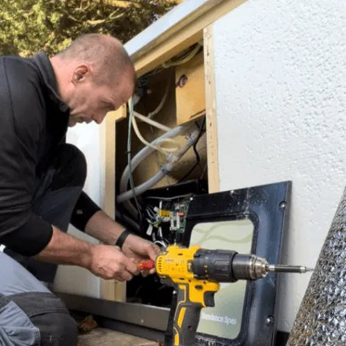 A Hot Tub Liverpool engineer wearing a black jacket and gray pants is kneeling beside an open panel of an outdoor spa, using a yellow electric drill and a red-handled tool to make adjustments to the internal components. Various hoses and wiring can be seen inside the panel during these Hot Tub Repairs.