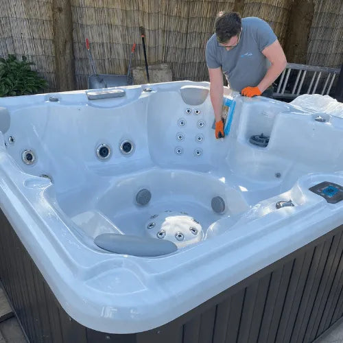 A person is performing hot tub maintenance on a large white tub with multiple jet nozzles, wearing an orange glove on one hand. They are applying a cleaning solution to the tub&#39;s surface. In the background, there is a bamboo fence and various gardening tools. This is part of their Hot Tub Servicing - Silver Package Clean and Check by Hot Tub Liverpool service.