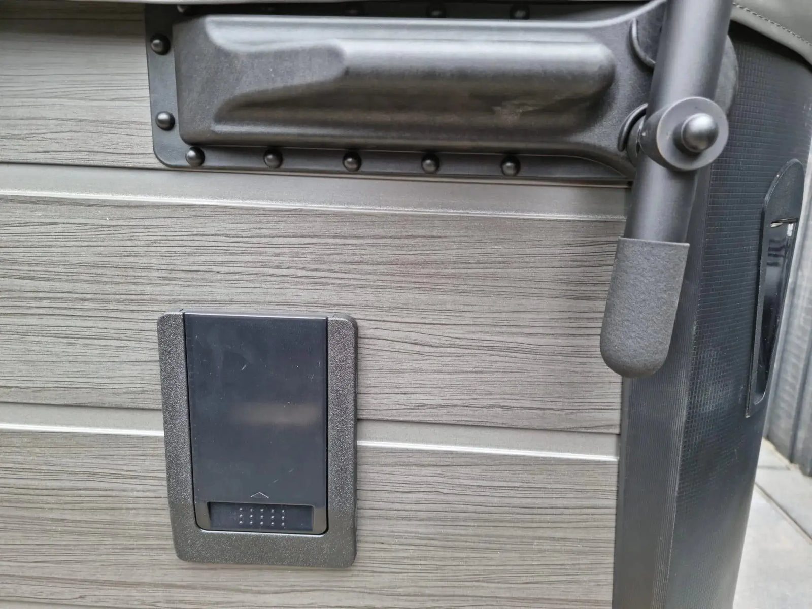 Close-up of a hot tub exterior featuring a handle, a rectangular panel, and the corner of a cover. The tub's surface has a wood-textured gray finish, and the panel is a small, black, door-like fixture. A section of an HTL Cover Lifter by Hot Tub Liverpool is visible on the right, showcasing practical hot tub accessories.