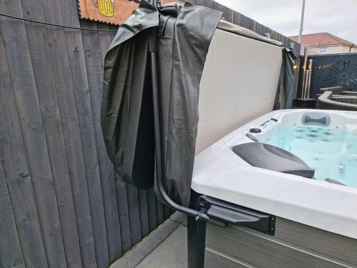 An outdoor hot tub with its cover partially lifted by a Hot Tub Liverpool HTL Cover Lifter, revealing the bubbling water inside. The hot tub is set against a dark wooden fence, and the cover rests on a metal stand to the side. The surrounding area includes a paved surface and additional fencing.