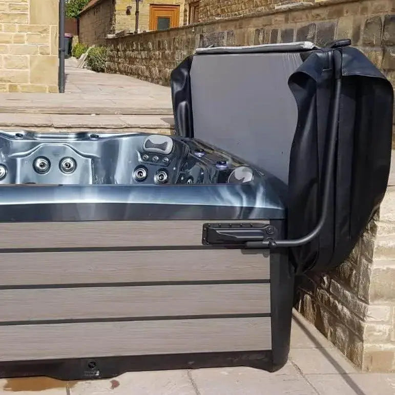 A hot tub with a gray and black exterior is situated outdoors next to a stone wall and sidewalk. The lid, aided by a Hot Tub Liverpool HTL Cover Lifter, is partially open, revealing multiple jets and seating inside the tub. In the background, there are buildings constructed of stone and brick.