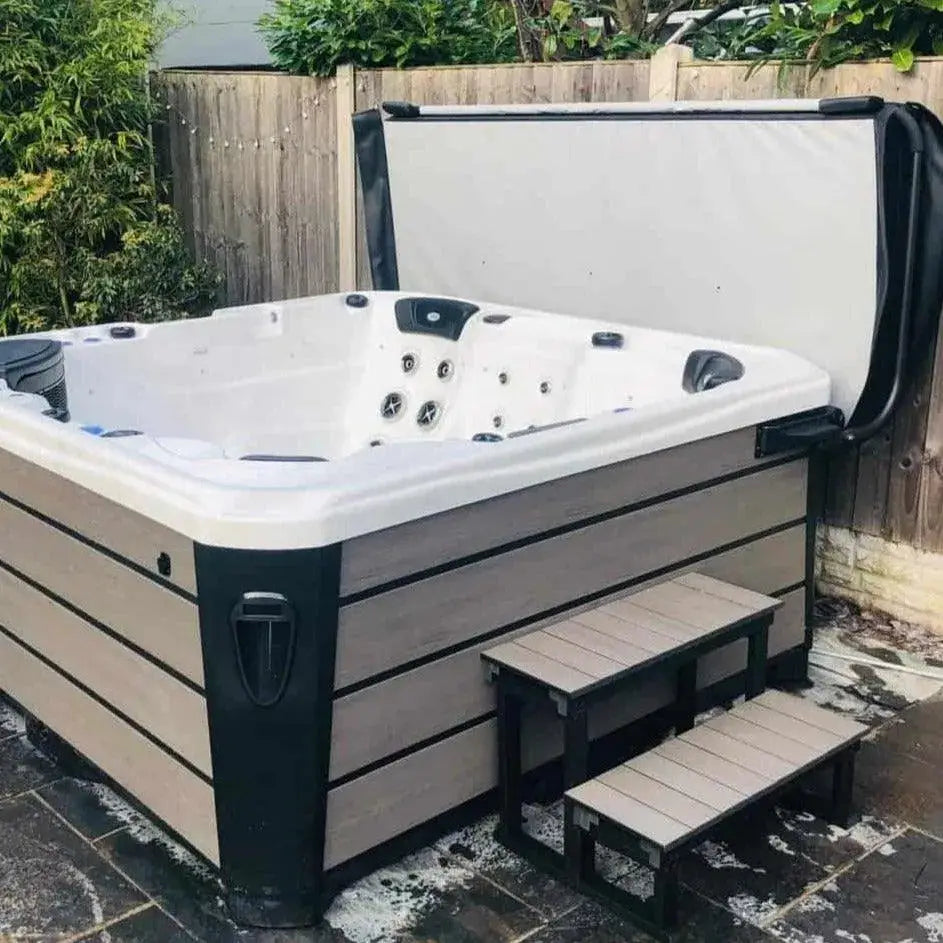 A large outdoor hot tub with a sleek design sits on a stone patio. It features multiple jet nozzles and an easy lifter for the hot tub cover, which is partially open and propped up by the HTL Cover Lifter from Hot Tub Liverpool. A small, matching two-step ladder provides easy access to the tub. Lush greenery and a wooden fence are in the background.