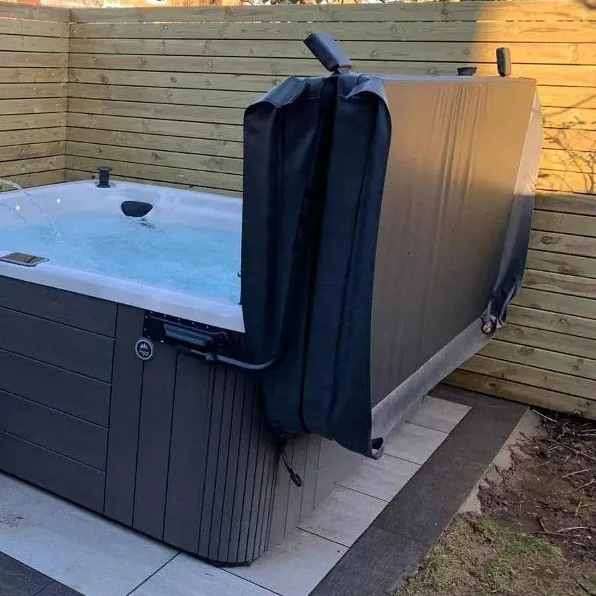 A hot tub with bubbling water is in a fenced backyard. The Hot Tub Liverpool HTL Cover Lifter has been used to remove the hot tub cover, which is propped up against one side. The surrounding area features a wooden fence and a paved section adjacent to the hot tub, showcasing the convenience of quality hot tub accessories.